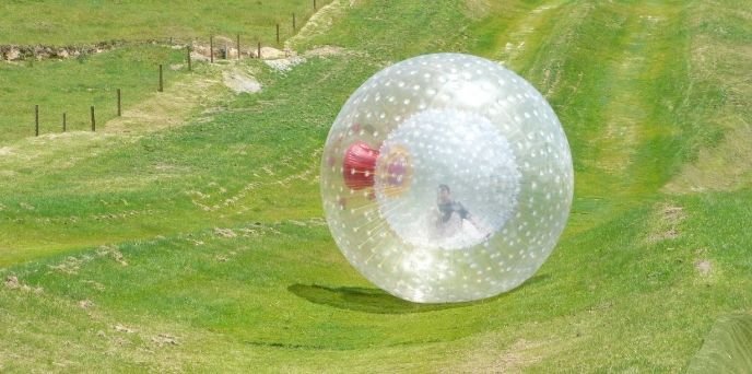 A person enjoys a zorbing adventure on a large inflatable ball, rolling down a hill surrounded by lush greenery in Manali.